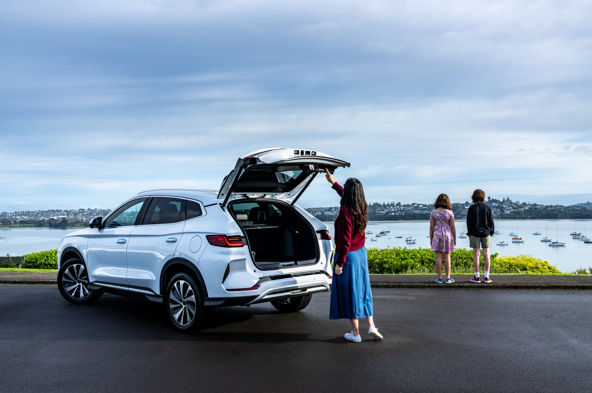 White BYD Sealion 6 Super Hybrid with lady closing rear door and two children standing, looking into bay with boats