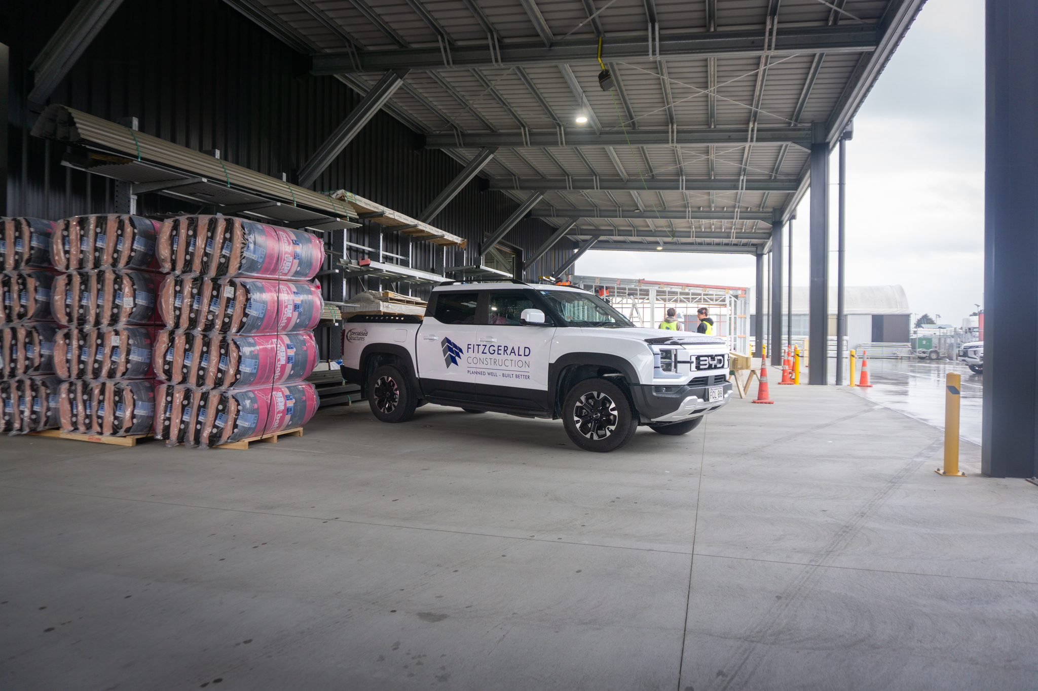 BYD Shark 6 with Fitzgerald Construction logo on side, parked at Fitzgerald Construction facility in Nelson. Pink Bats insulation bales on side with house construction behind.